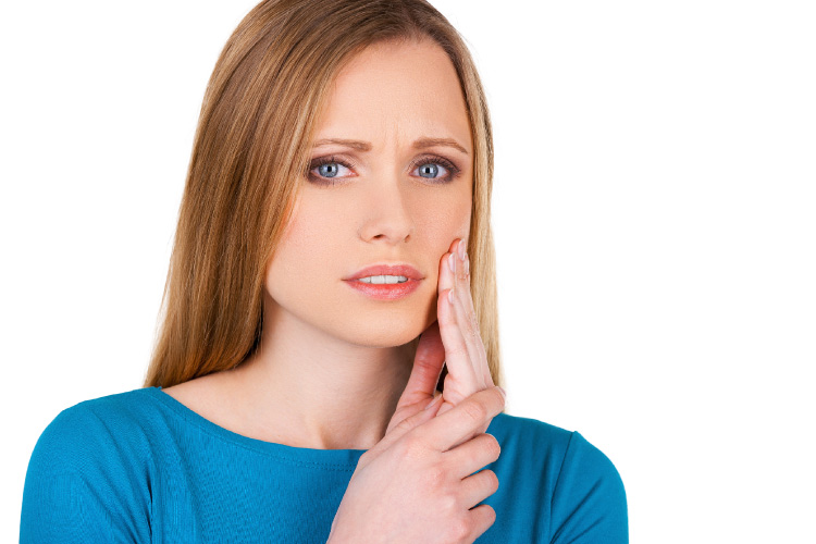 Blonde woman in a blue shirt cringes and touches her cheek due to sensitivity and tooth pain