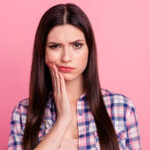 Brunette woman frowns in pain and touches her cheek due to a dental emergency against a pink wall