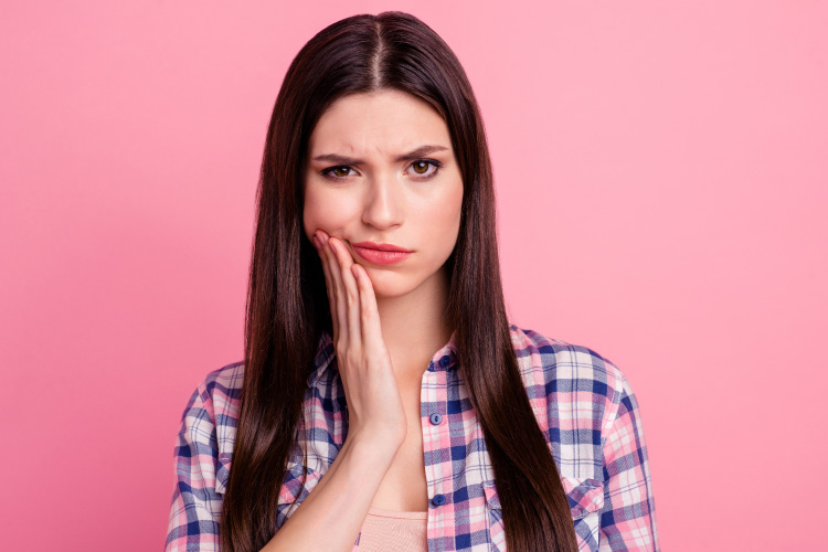 Brunette woman frowns in pain and touches her cheek due to a dental emergency against a pink wall