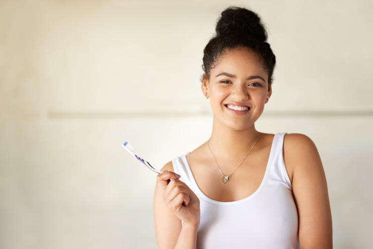 Young woman with dark hair and a white tank top smiles while holding a toothbrush to care for her oral hygiene during COVID-19