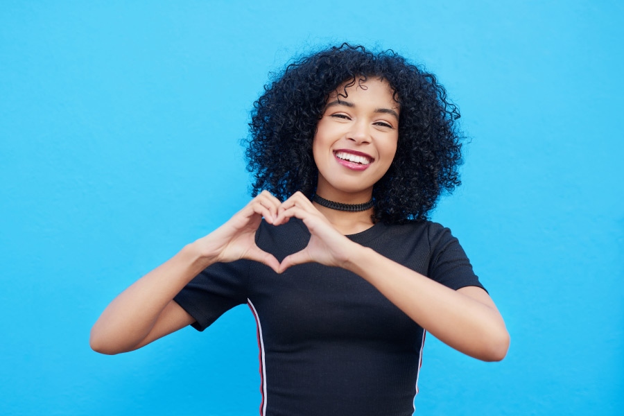 Curly-haired woman in a black shirt against a blue wall smiles and makes a heart shape for Optima Dental Spa