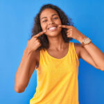 Brunette woman in a yellow tanktop smiles and points at her teeth after her cosmetic consultation