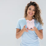 Curly-haired woman in a blue shirt smiles as she holds a piggy bank to discuss Optima Dental Spa's Dental Savers Plan
